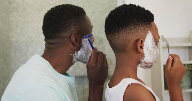 African American father and son shaving beards together in bathroom, demonstrating bonding moment and childhood learning skill. Ideal for family, parenting, and grooming concept. Use in advertisements for shaving products, family lifestyle blogs, or social media campaigns celebrating fatherhood.