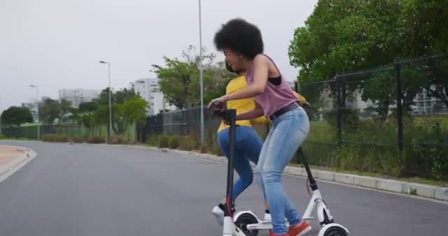 These biracial women are having fun together, riding electric scooters in a city park. Great for promoting urban lifestyle, modern transportation options, outdoor activities, and leisure time with friends. Ideal for use in urban planning materials, advertisements for electric scooters, travel promotions, and social media posts highlighting outdoor fun.