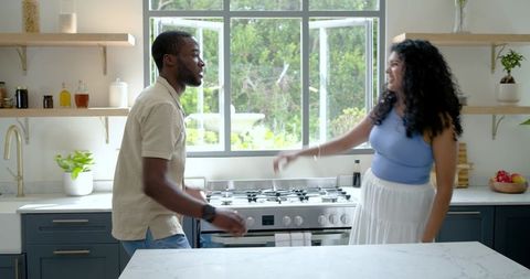 Sunlit diverse couple cooking together in modern kitchen with marble island and stove