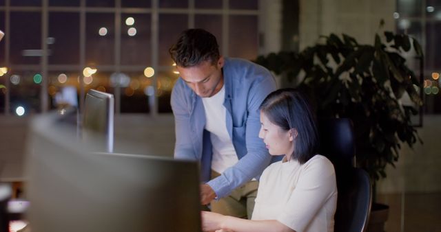 Professionals are collaborating over computer screens in a dimly lit office during late hours. Ideal for articles on work culture, teamwork, office settings, technology usage in business, overworking environments, and corporate teamwork scenarios.