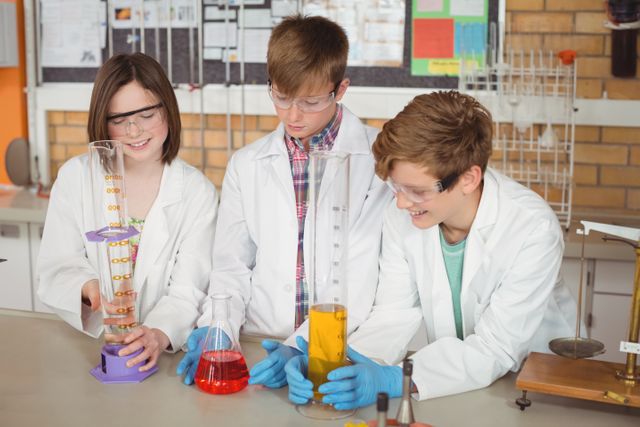 Three schoolchildren wearing lab coats and safety goggles are conducting a chemical experiment in a school laboratory. They are handling beakers and measuring cylinders filled with colorful liquids. This image is ideal for educational content, science-related articles, and promotional materials for STEM programs.