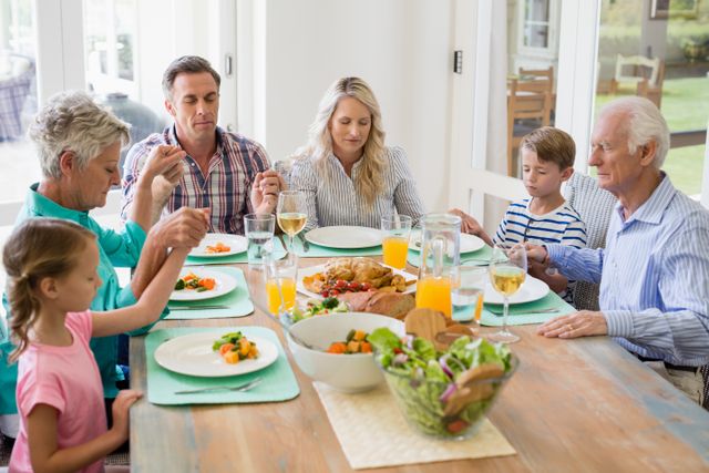 Multi-generation family holding hands and praying before a meal at home. Grandparents, parents, and children gathered around a dining table with a variety of food and drinks. Ideal for use in articles or advertisements about family values, traditions, and togetherness.