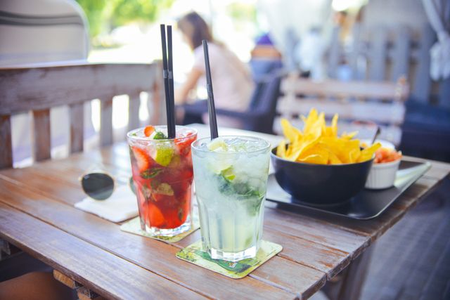 Summer cocktails with mojito and strawberry cocktail on a wooden table at an outdoor café. A bowl of nachos serves as a perfect snack pairing. Great for showcasing summer refreshments, outdoor dining, leisure activities, and relaxation. Ideal for marketing food and beverage promotions, cafe advertisements, and lifestyle content.