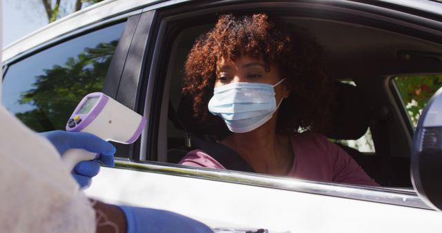 African american woman with face mask sitting in car having temperature measured by medical worker. health checks and precautions during coronavirus covid 19 pandemic.