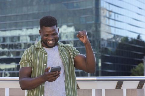 African american man celebrating with smartphone on urban terrace balcony by glass office