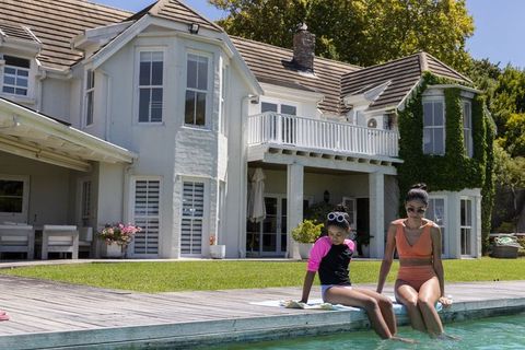 African american mother and daughter relaxing by pool, dipping toes in sunlit backyard