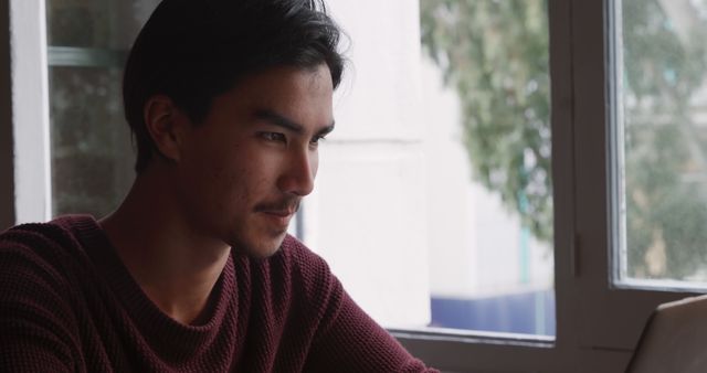 Young man concentrates while working on laptop indoors next to window. The natural light adds to the focused atmosphere. Perfect for depicting remote work, studying, office environments, or digital nomad lifestyle.