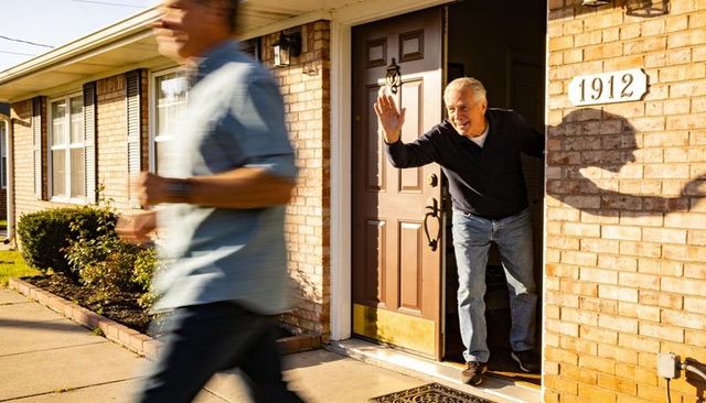 Senior man waving from front door leaning out to greet neighbor on sunny suburban porch