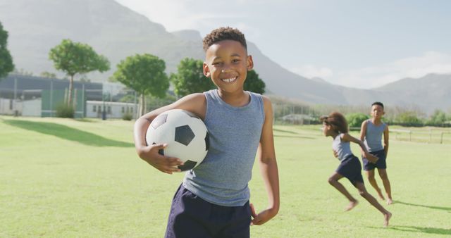 Young boy holding a soccer ball and smiling while standing on a green grass field under a sunny sky. Other children playing in the background. Great for content related to sports, outdoor activities, children's health, teamwork, and active lifestyle.