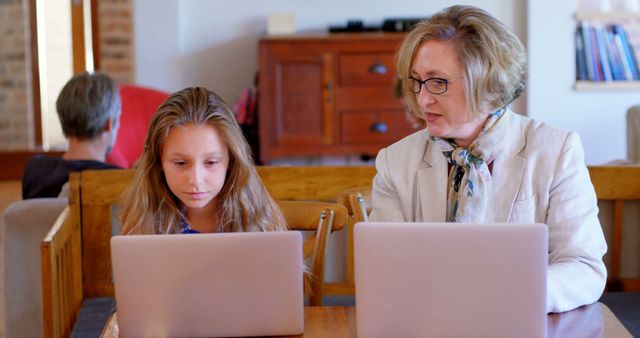 This image depicts an older woman and a young girl working on laptops at a domestic table. Ideal for use in articles or advertisements related to online education, family tech usage, remote work, homeschooling, generational learning, and parental supervision of digital activities.