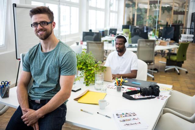 Portrait of male graphic designer smiling at camera while colleague working at desk in background