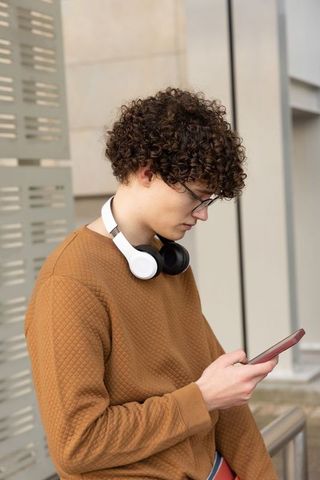 Teen male checking smartphone while standing outdoors wearing headphones and glasses