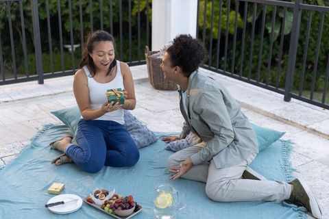 Woman in 20s receiving surprise gift from man in 30s during terrace picnic with fruit