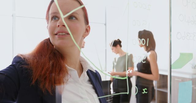 Businesswoman presenting ideas on glass board in modern office with coworkers discussing in background. Image can be used for business presentations, teamwork and collaboration projects, corporate strategies, and office environments showcasing professional teamwork.