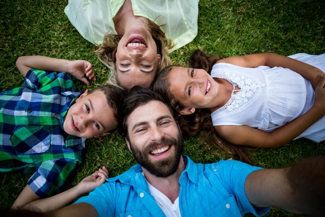 This image shows a cheerful family of four lying on grass, smiling at the camera. Ideal for use in advertisements, family-oriented content, lifestyle blogs, and promotional materials for outdoor activities or family events. It conveys themes of happiness, togetherness, and outdoor fun.