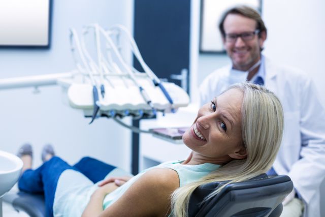 Female patient smiling while sitting in a dental chair with a dentist in the background. Ideal for use in healthcare, dental care, and medical service promotions. Suitable for illustrating dental checkups, professional dental services, and patient satisfaction.