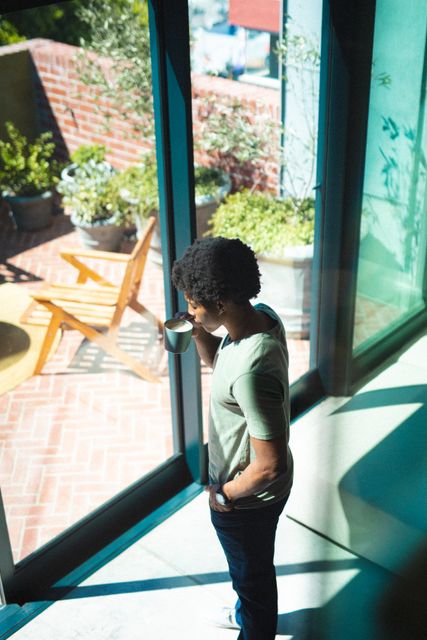 Young African American man stands by a sliding window, drinking coffee and looking outside. The scene captures a moment of contemplation and relaxation in a modern home setting. Ideal for use in lifestyle blogs, articles on morning routines, advertisements for coffee brands, or content related to modern living and home decor.