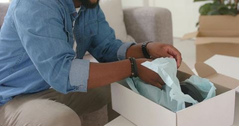 African man unpacking delivery box at home revealing headphones wrapped in blue tissue