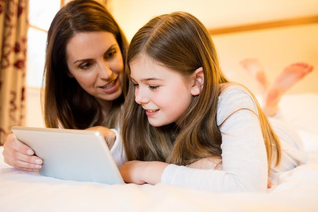 This image shows a mother and daughter lying on a bed using a tablet. They are smiling and seem engaged with the device, indicating a moment of family bonding and shared enjoyment. Ideal for use in articles or advertisements about family activities, parenting tips, technology in education, or home life. It could also be used in content promoting digital literacy and family-oriented technology products.