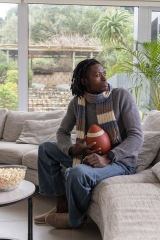 African american man relaxing on sofa holding football watching game with popcorn