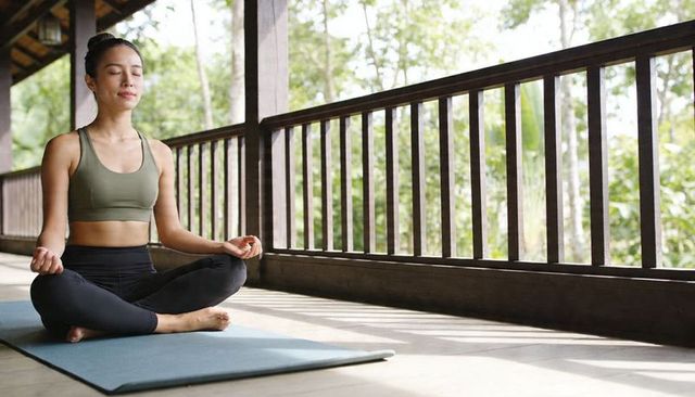 Asian woman meditating on wooden veranda practicing yoga and mindfulness outdoors at sunrise