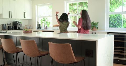 Two friends chatting at modern bright kitchen island with leather barstools and greenery