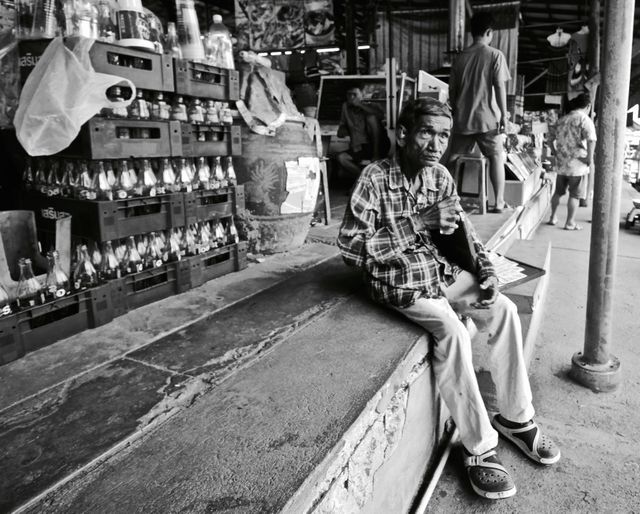 This black and white image captures an elderly man sitting on stairs in a traditional market. He appears thoughtful while holding a drink; the market backdrop features stacked soda bottles and other patrons in the background. Ideal for illustrating storytelling elements involving daily life, market culture, elderly populations, and community interactions. It can be used for editorial purposes, blogs highlighting traditional markets, or to provide a slice-of-life context in various multimedia projects.
