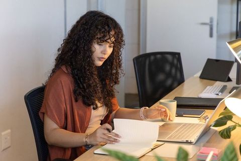 Curly-haired woman working on laptop and paperwork at organized coworking desk with coffee mug