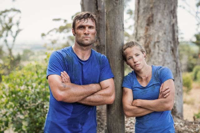 Man and woman standing with arms crossed, appearing tired and sweaty, during an outdoor obstacle course in a boot camp. Ideal for use in fitness, training, and teamwork-related content, as well as advertisements for outdoor activities and endurance challenges.