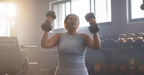 Mid-adult woman performing standing dumbbell overhead press in gym with kettlebells behind