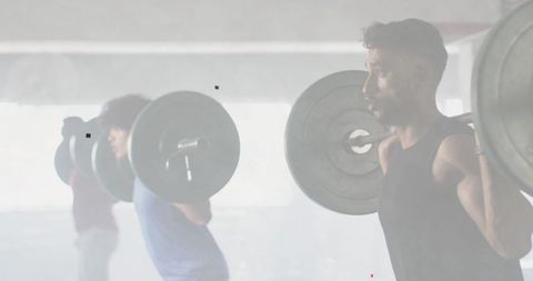 Backlit man squatting with loaded barbell in industrial gym during group weightlifting