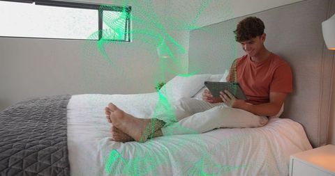 Young man relaxing on bed with tablet in modern minimal bedroom, smiling, daylight
