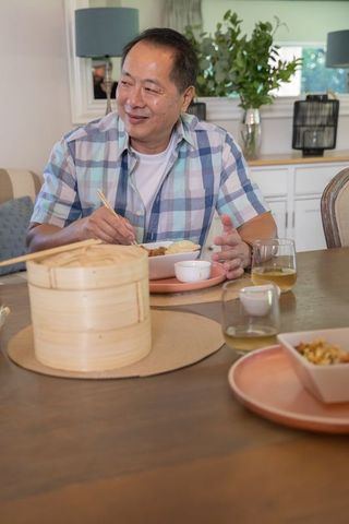 Senior asian man enjoying casual home dinner using chopsticks near bamboo steamer