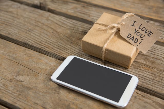 This image shows a gift box with a tag that reads 'I Love You Dad' next to a smartphone on a wooden table. It is perfect for use in Father's Day promotions, greeting cards, social media posts, and advertisements celebrating fathers and expressing love and appreciation. The rustic wooden table adds a warm, homely feel to the scene.