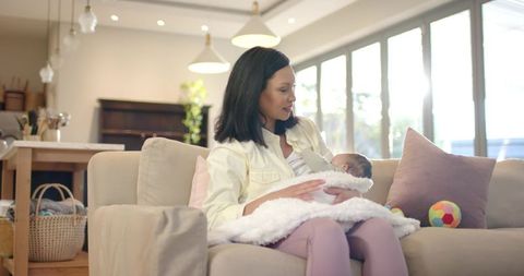 Asian Mother Feeding Newborn on Comfortable Sofa in Sunlit Living Room