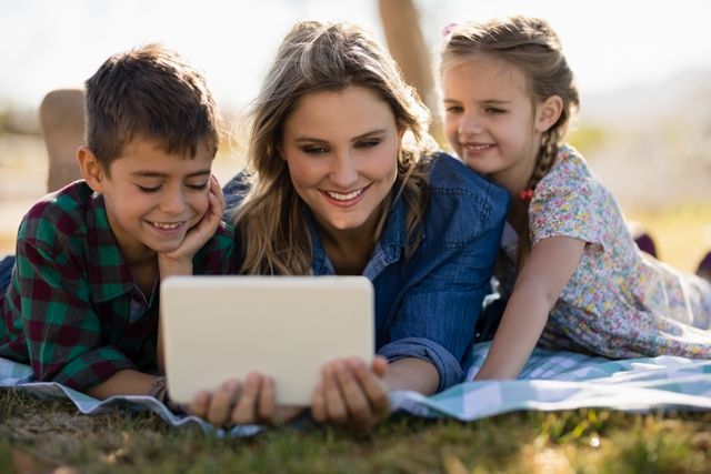 Mother and two children lying on a blanket in a park, smiling and looking at a digital tablet. Ideal for promoting family bonding, outdoor activities, technology use in family settings, and leisure time. Suitable for advertisements, blogs, and articles about parenting, family life, and digital learning.