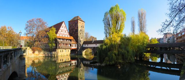 Medieval architecture and half-timbered house reflecting in calm river with willow trees and autumn colors. Ideal for travel blogs, tourism brochures, and content showcasing historical landmarks. Highlights tranquility and scenic views of old city landscapes.