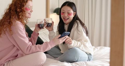 Two friends enjoying cozy spa night with under-eye patches, hot drinks and smartphone