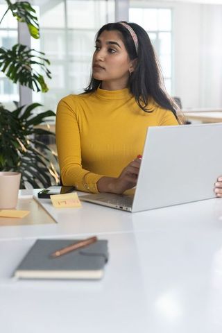 Indian professional woman working on laptop in modern office wearing mustard turtleneck