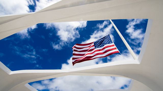 An American flag flying at the Pearl Harbor Remembrance Memorial against a bright blue sky with white clouds. Useful for themes related to patriotism, U.S history, national holidays, veterans, and remembrance events. Suitable for educational materials, patriotic campaigns, and historical documentaries.