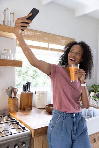 Young woman taking selfie while holding orange juice, smiling casual morning kitchen routine