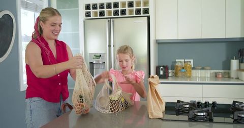 Mother and daughter unpacking groceries with reusable mesh bags in sunlit modern kitchen