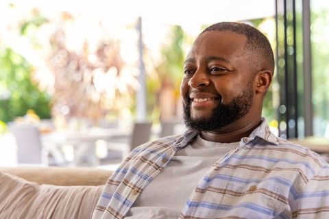 Relaxed man smiling on sunlit sofa near glass doors, casual home interior lifestyle