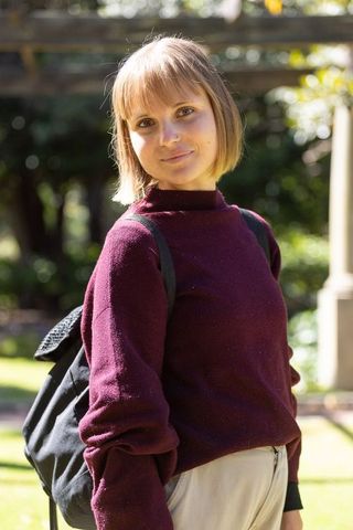 Young female student standing on campus green wearing maroon sweater and backpack, sunlit