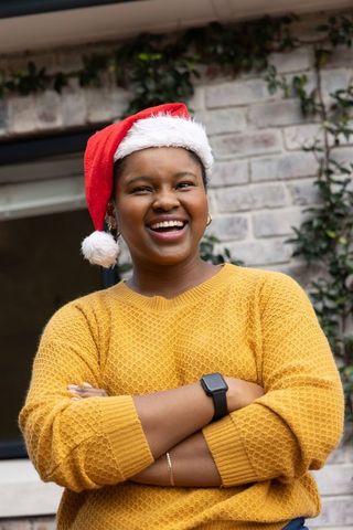 African american woman smiling wearing santa hat mustard sweater and smartwatch outdoors