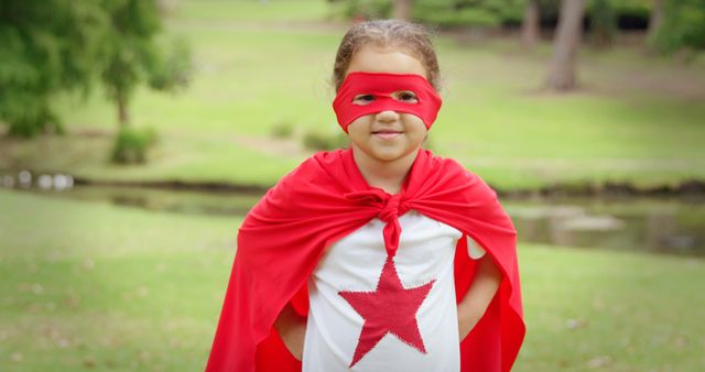 Young girl in superhero costume with red cape and mask stands confidently in a park. Perfect for concepts related to childhood imagination, play, creativity, and empowerment. Can be used in educational materials, advertisements promoting children's products, or content inspiring bravery and confidence.