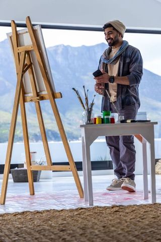 Smiling artist in beanie painting in bright studio with mountain view and art supplies