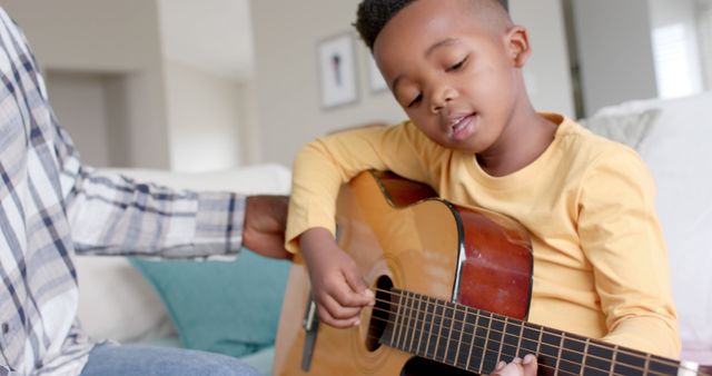 A young boy focused on playing an acoustic guitar, sitting comfortably on a couch in a cozy living room. An adult hand, likely a father's, assists from beside. The image captures a moment of family bonding, music practice, and home life. Ideal for illustrating concepts related to music education, parent-child relationships, and home leisure activities.