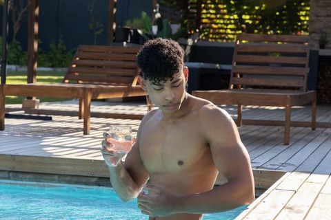 African american man relaxing in backyard pool holding glass of water on sunlit deck