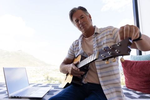Senior man tuning acoustic guitar on daybed by window with laptop and panoramic view